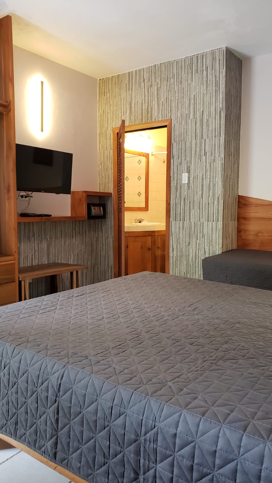 Hotel room with bed, TV, and open bathroom door. Gray quilted comforter. Gray textured wall.