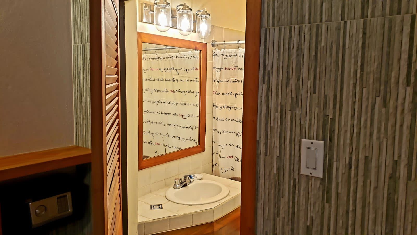Bathroom with sink, mirror, and shower, viewed through an open doorway. Stone wall texture on right.