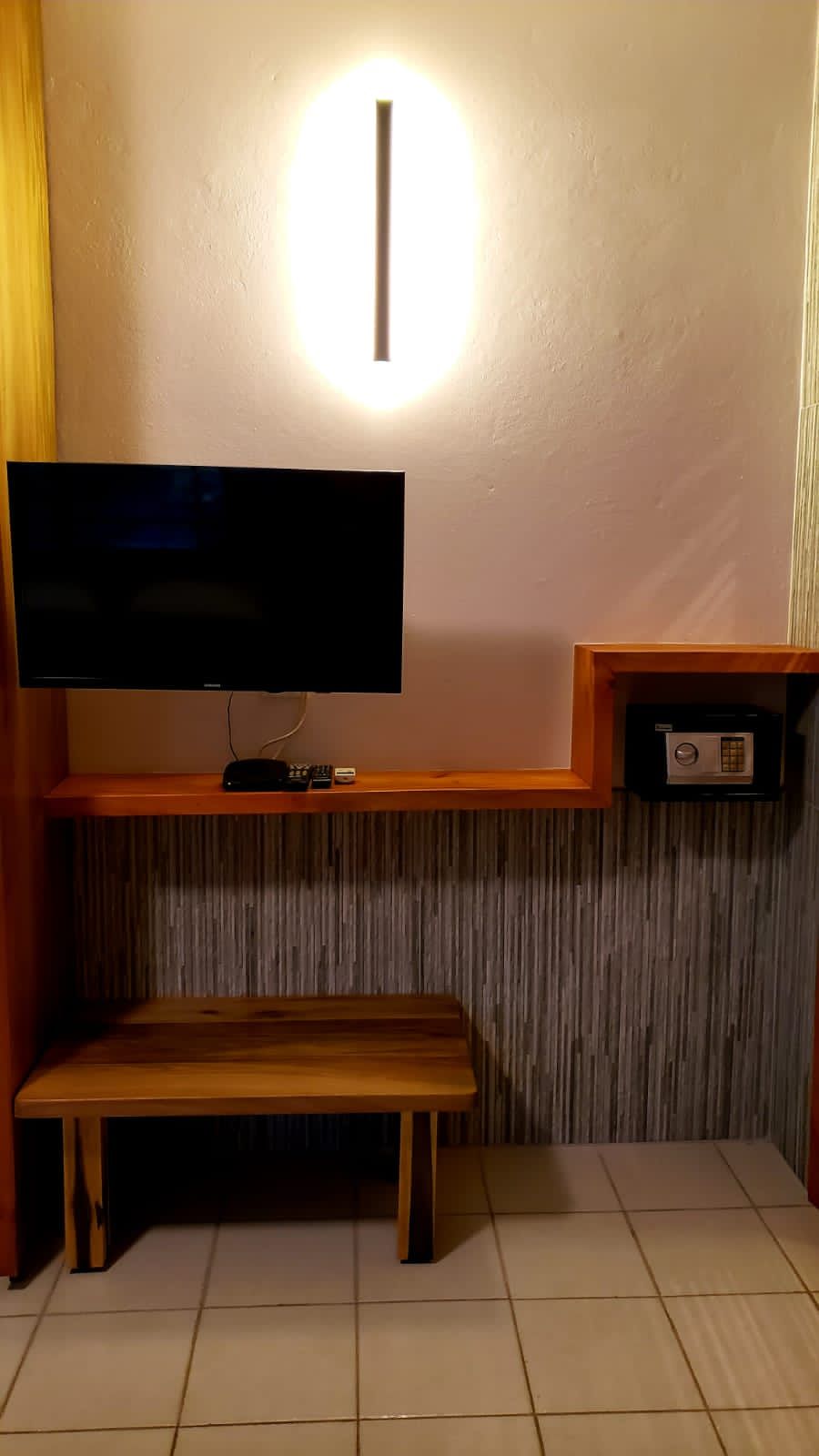 Hotel room wall with mounted TV, shelf with safe, bench, and strip lighting.