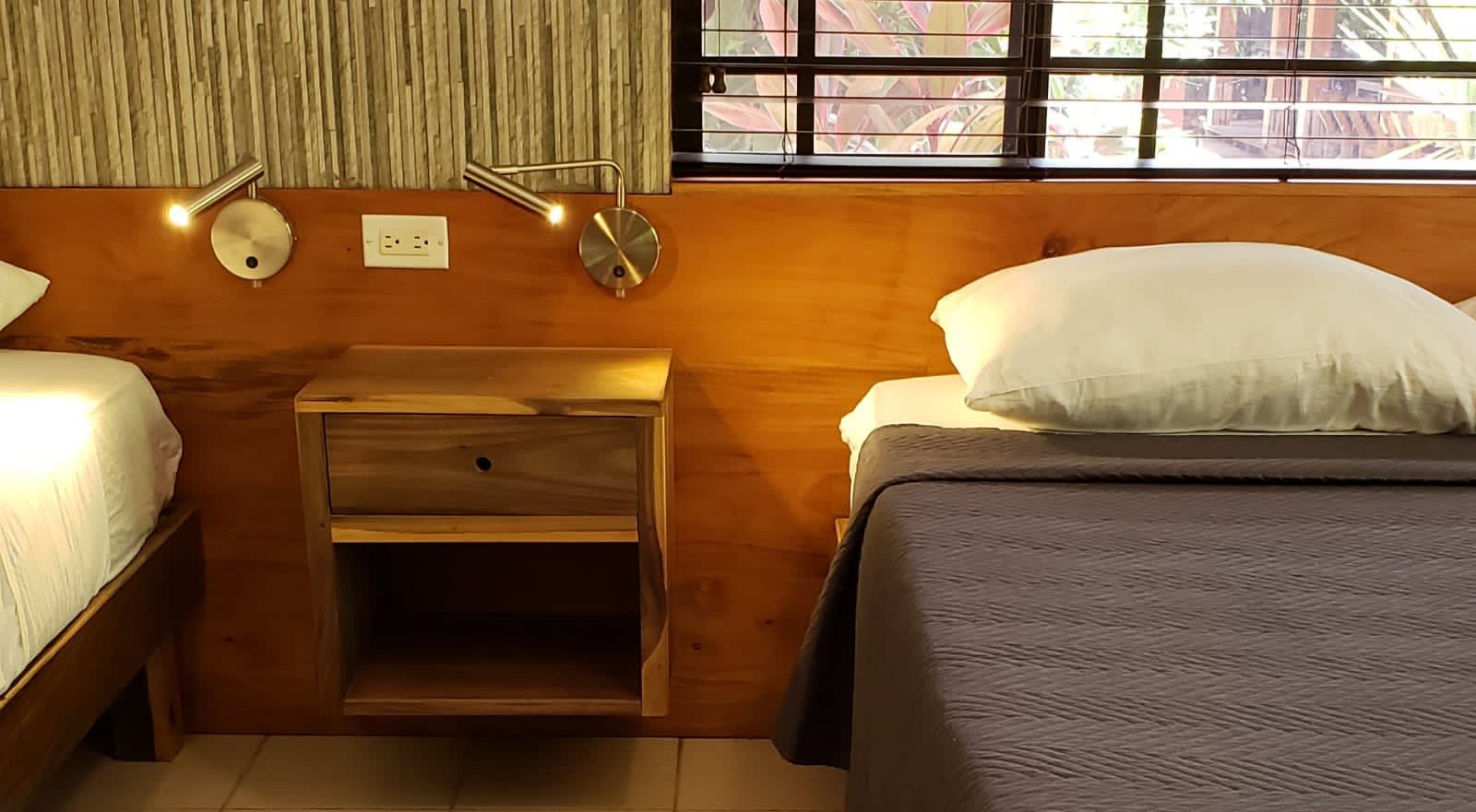 Two beds in a room with a wooden headboard, nightstand, and window. Gray and white bedding.