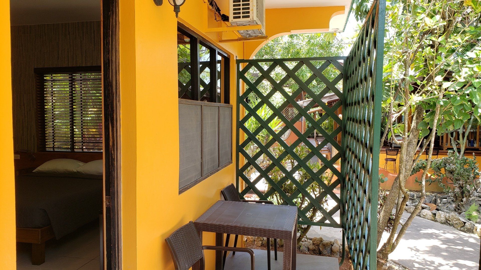 Yellow-walled patio with small table and chair, lattice screen, and partial view of a bedroom interior.