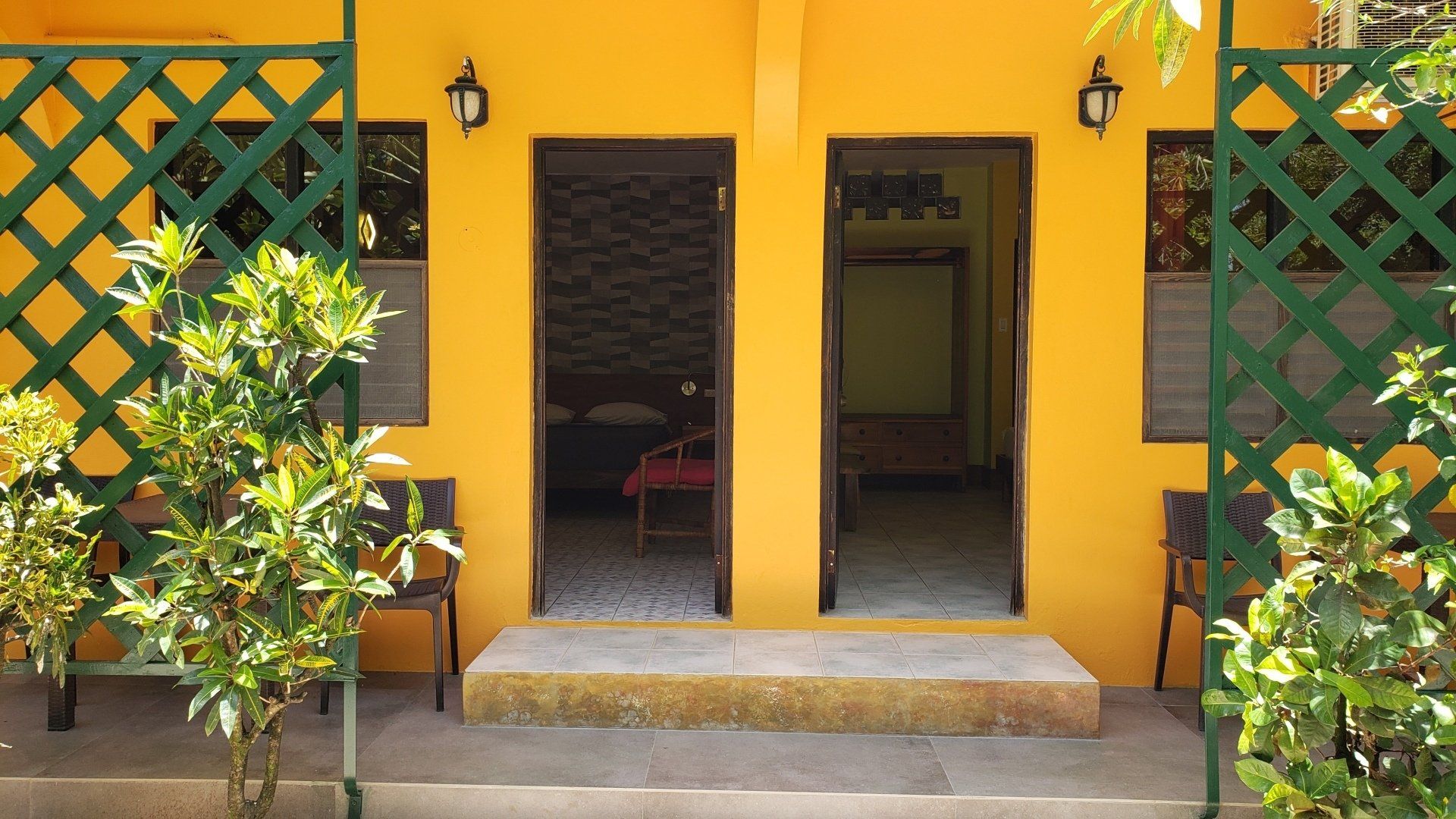 Yellow building with two open doorways, green trellis, plants, and concrete steps.