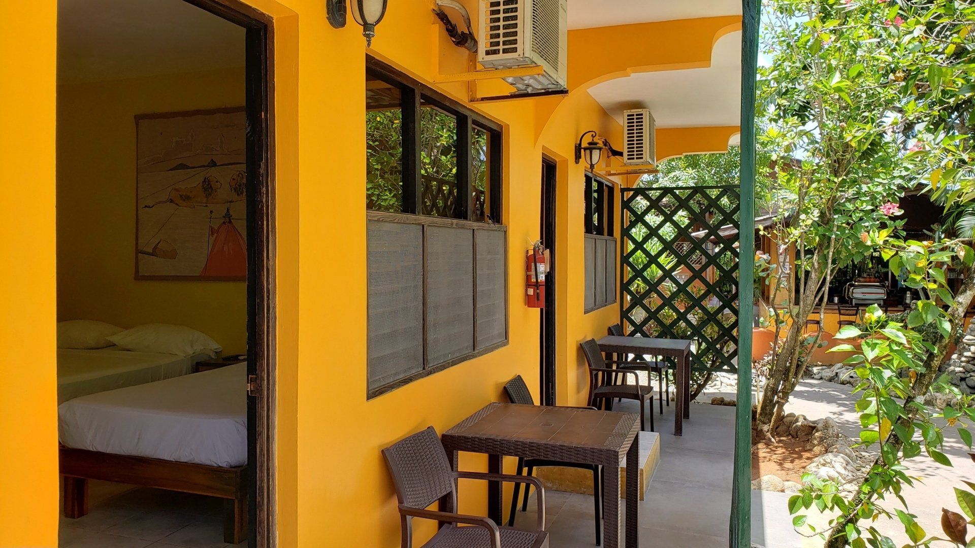 Yellow-painted building exterior with a shaded seating area, door to a bedroom, and lush greenery.