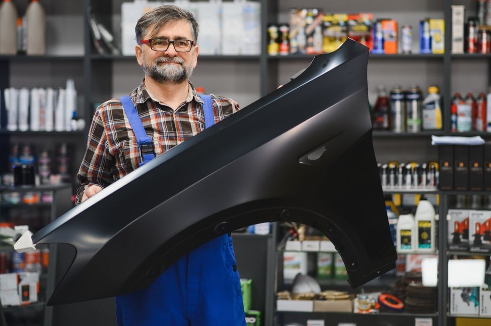 Man in a workshop holding a large black car body panel in front of shelves of paint cans