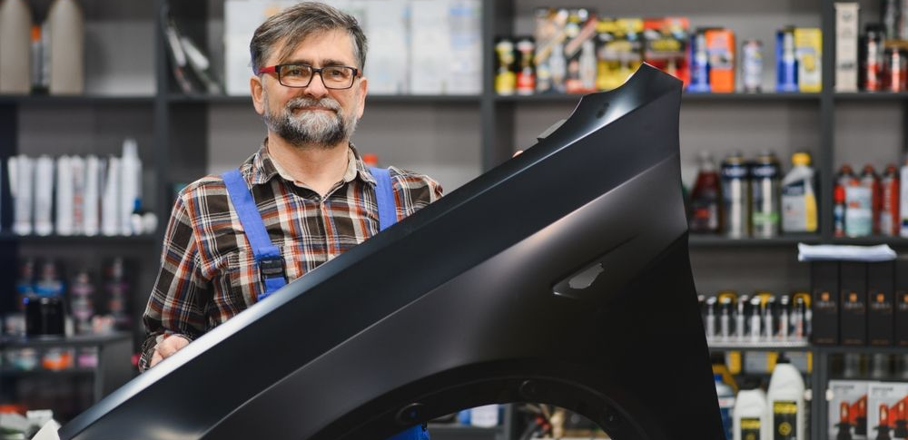 Mechanic smiling behind a car hood in an auto repair shop with shelves of supplies