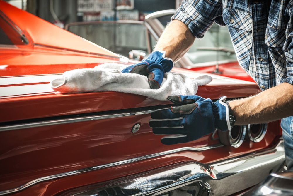Gloved hands polishing a red classic car with a white cloth in a garage