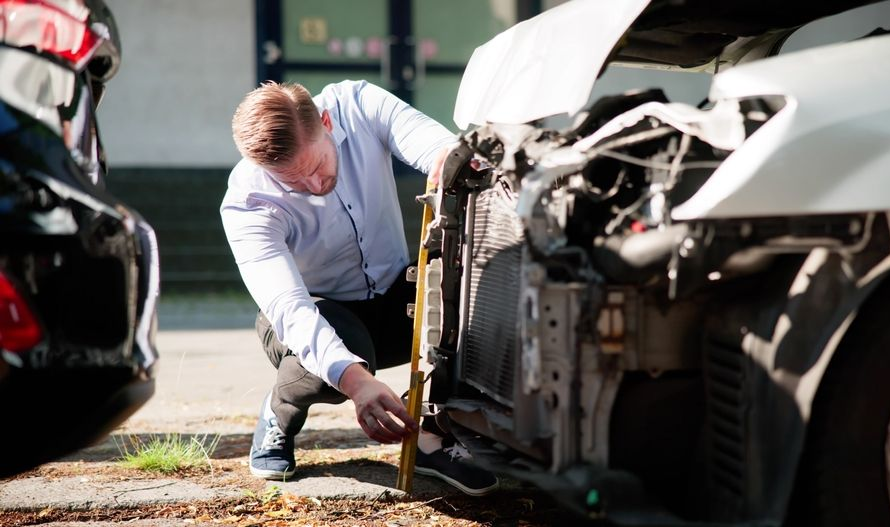 Mechanic inspecting damage on a white car after a crash, with a damaged vehicle in the foreground.