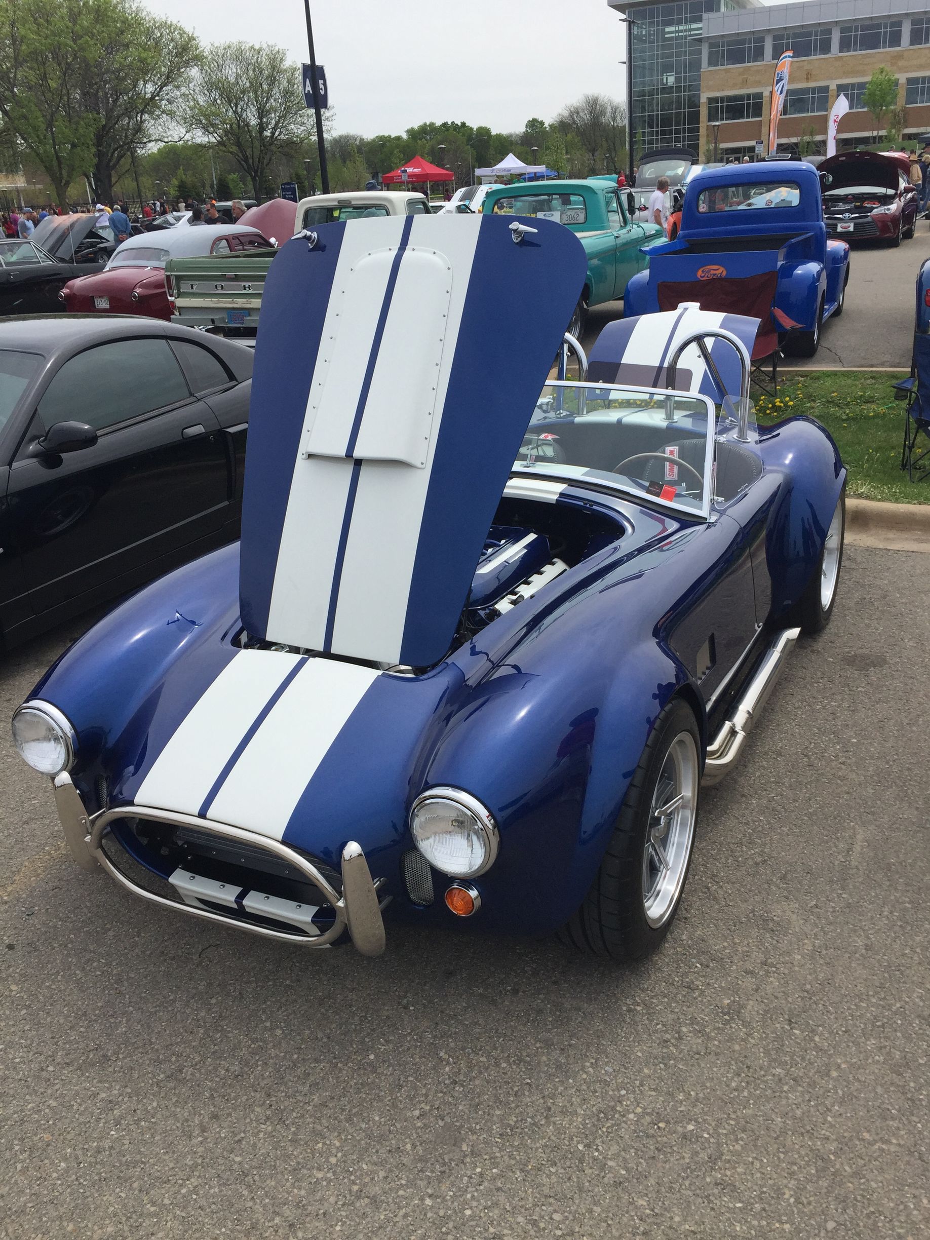 Blue classic roadster with white racing stripes displayed at a car show with hood open for viewing.