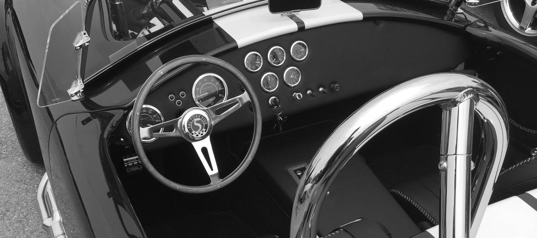 Black-and-white interior view of a classic roadster showing steering wheel, gauges, and chrome roll bar.