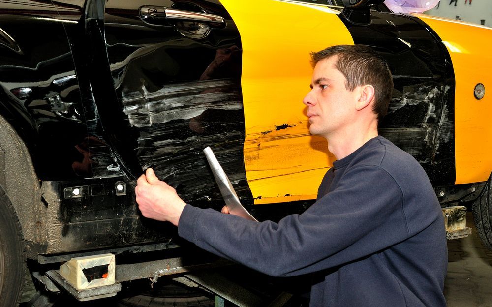 Mechanic inspecting a black and yellow car's damaged side panel in a garage