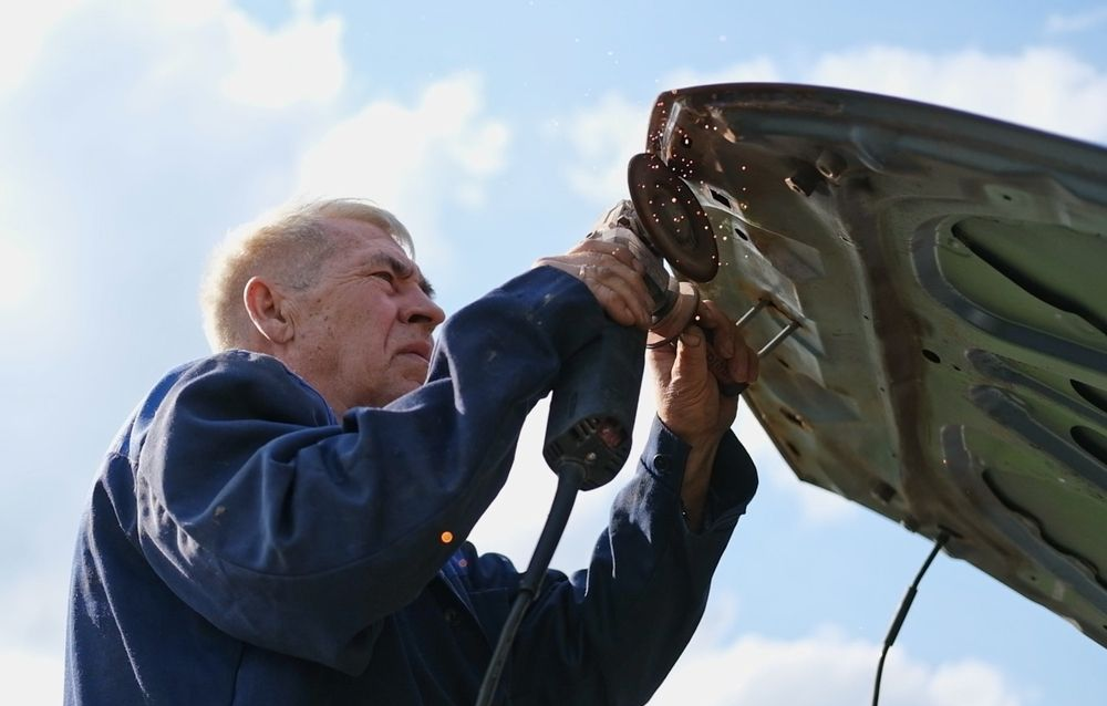 Man in blue jacket inspecting or repairing the underside of a large vehicle against a blue sky