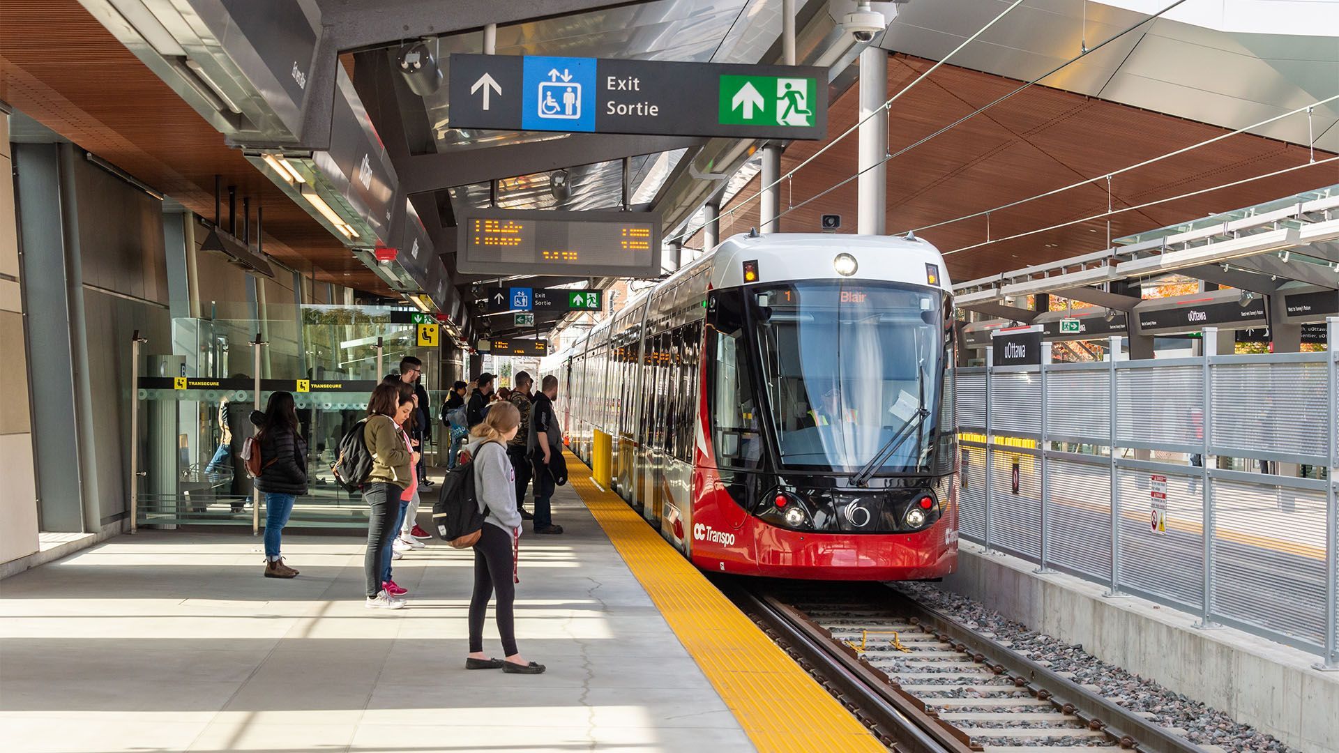 Modern tram at a covered station platform with waiting passengers and overhead signs