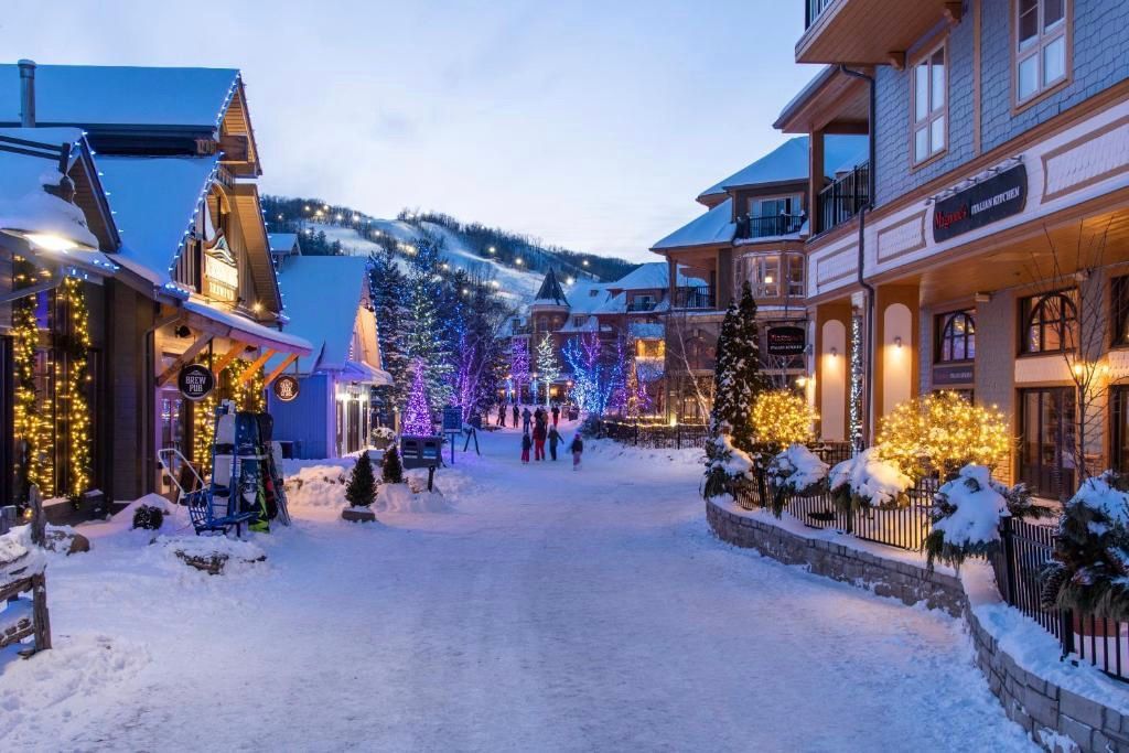 Snow-covered ski village street at dusk, lined with lit shops and chalets under a blue winter sky