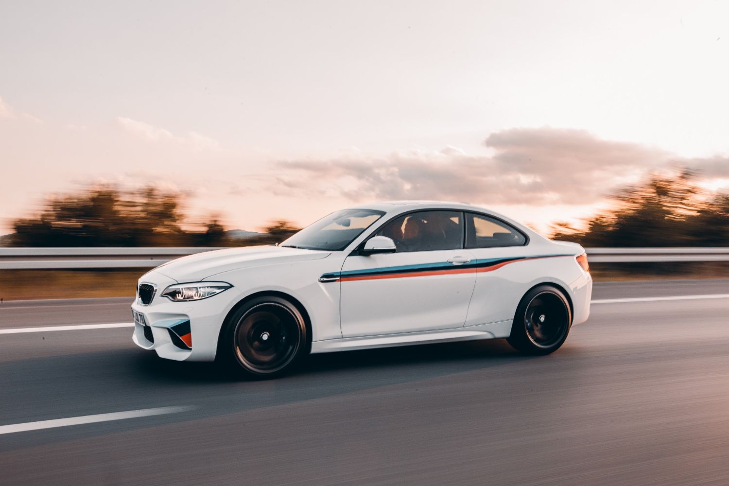 A white bmw m2 is driving down a highway at sunset.