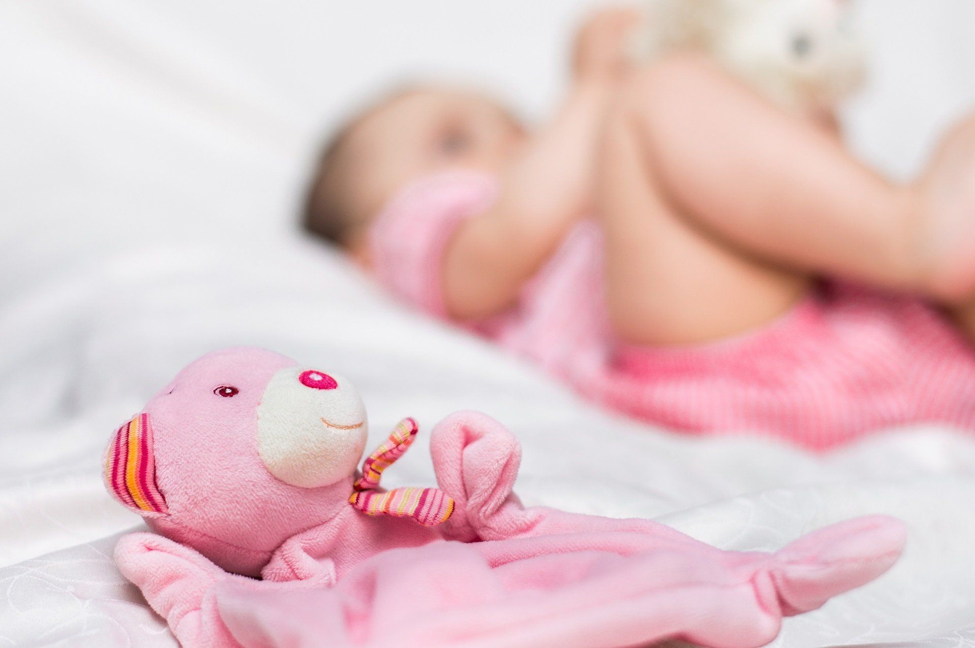 Pink teddy bear lying on a white blanket, baby in pink outfit in soft focus in the background.