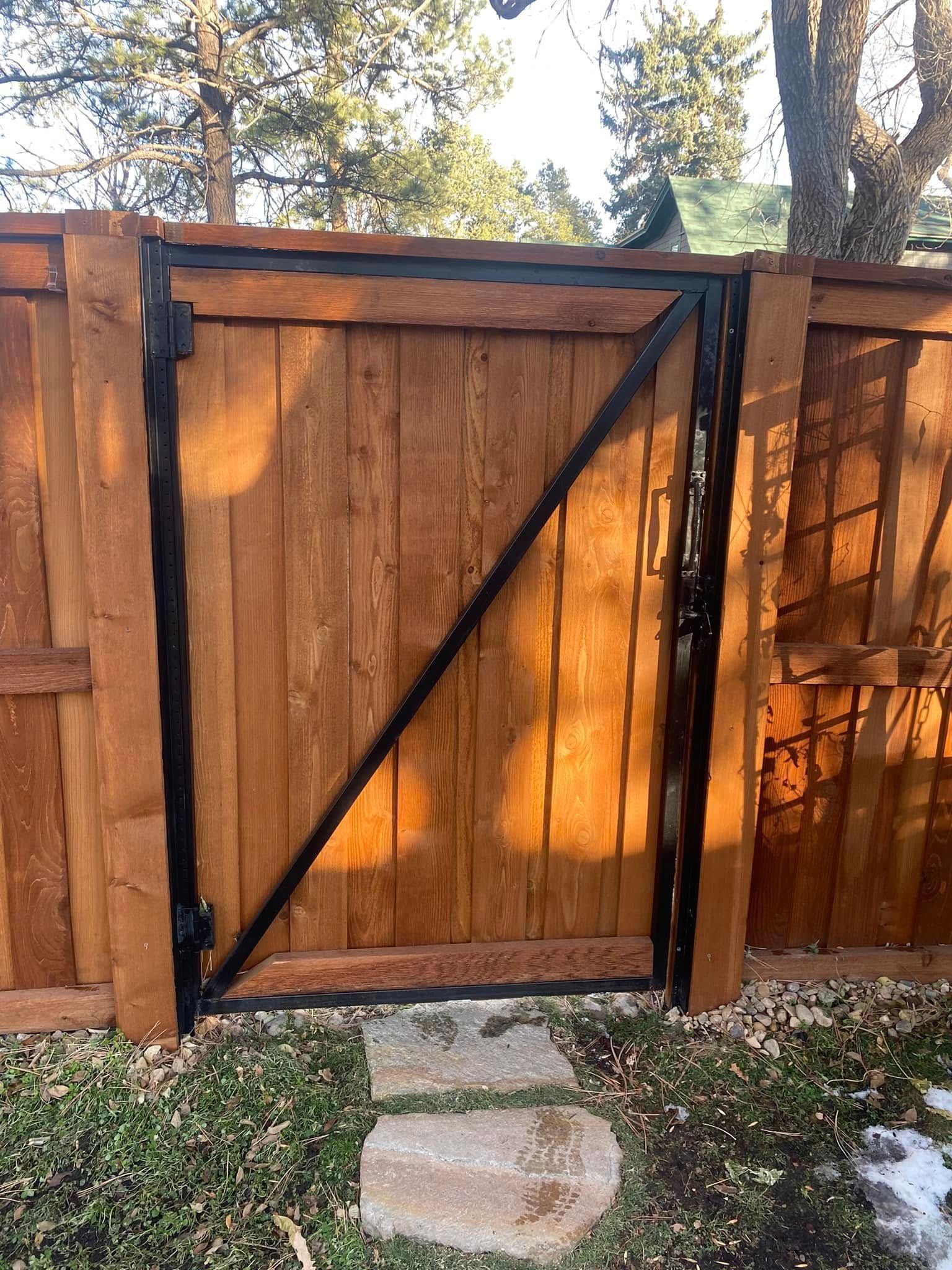Wooden fence gate with black diagonal bracing, sunlit in a backyard beside grass and stone path.