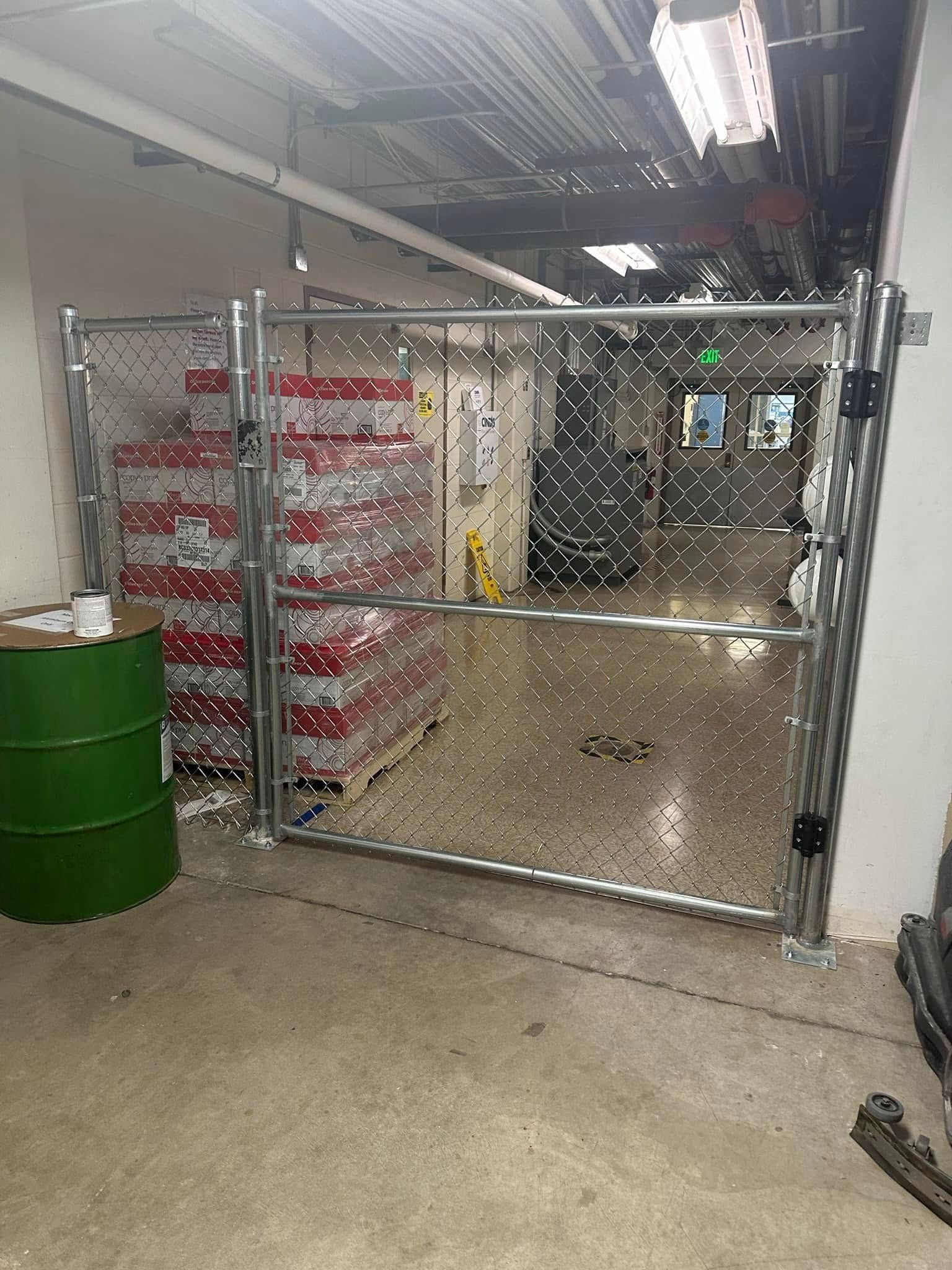 Chain-link gate in an industrial hallway, with red stacked bins on the left and a green barrel in front.