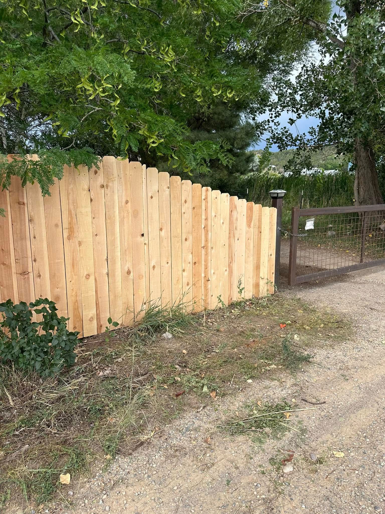 Wooden fence along a dirt path beside green trees and brush, with a metal gate in the background.