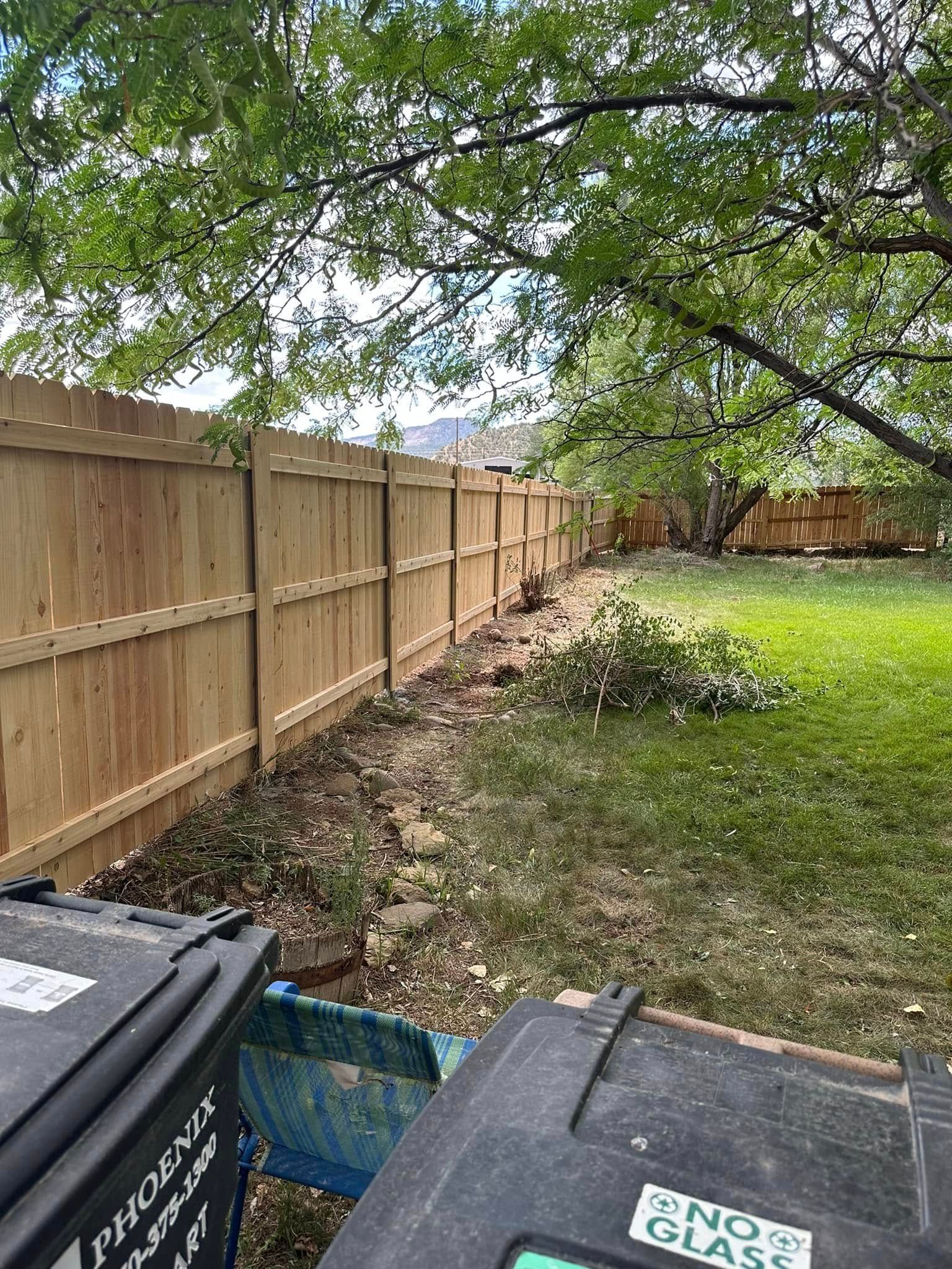 Backyard with wooden fence, leafy tree canopy, grass, and trash bins in the foreground