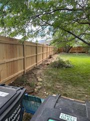 Backyard with wooden fence, leafy tree canopy, grass, and trash bins in the foreground