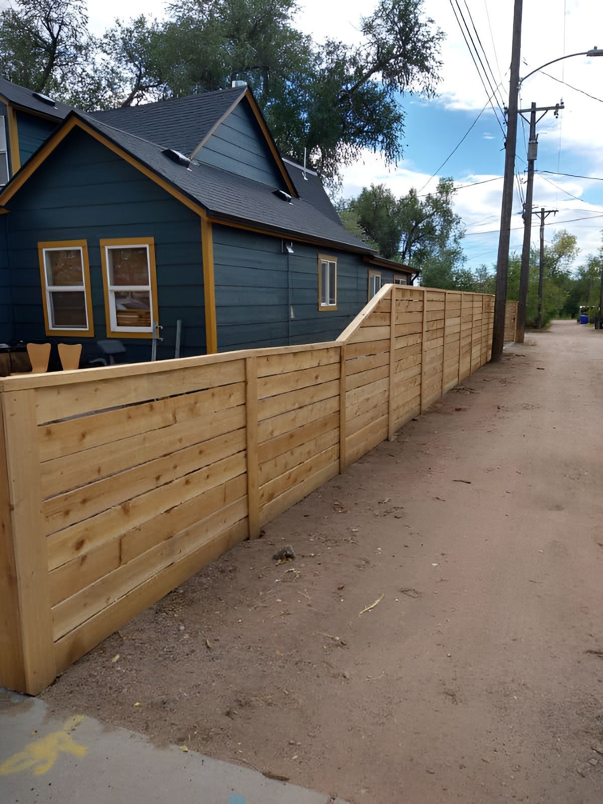 Long wooden fence beside a dark blue house along a dirt road under a cloudy sky