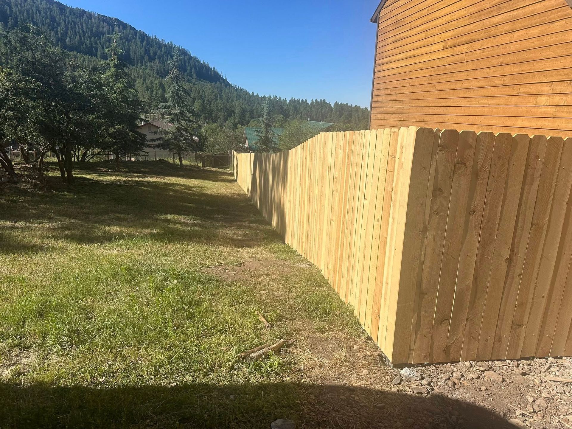 Wooden fence along a grassy yard beside a brick house, with trees and hills in the background.