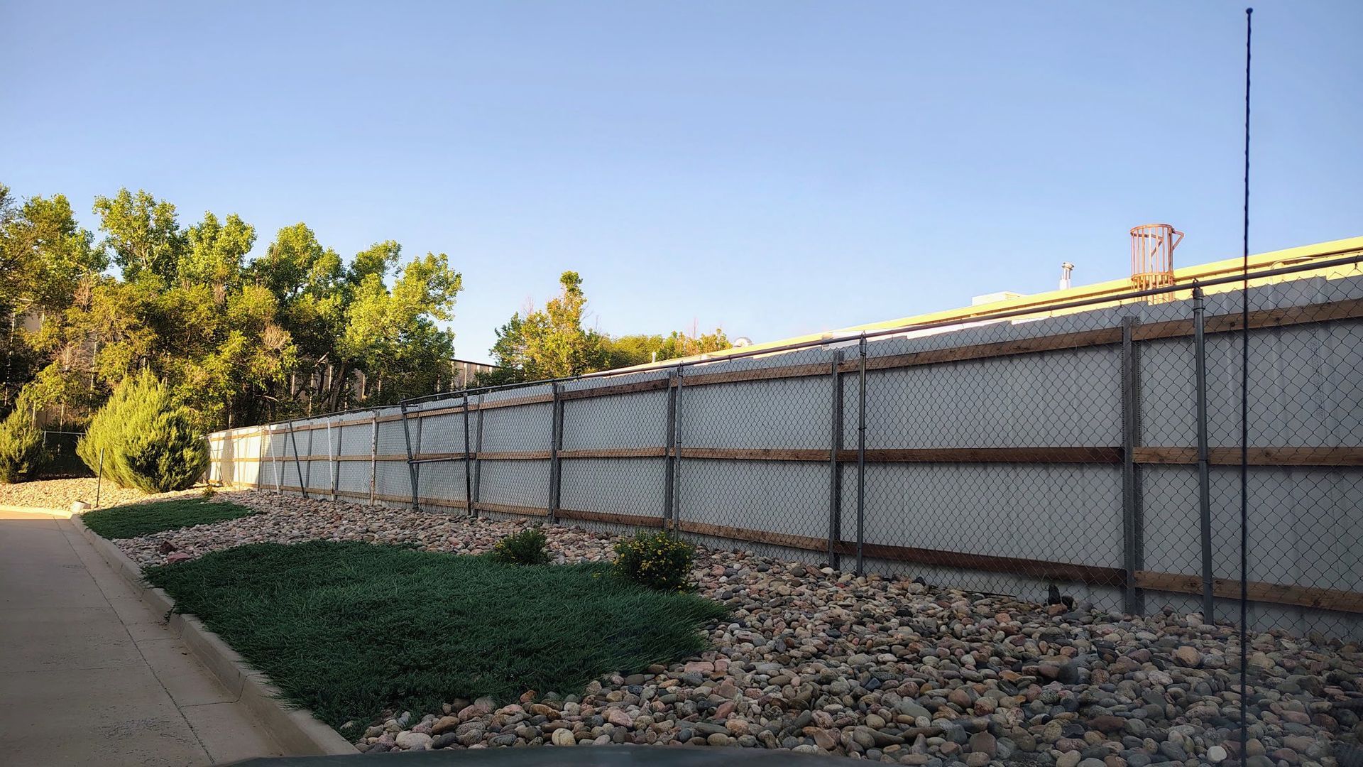Long fence beside a sidewalk and gravel strip, with trees under a clear blue sky.