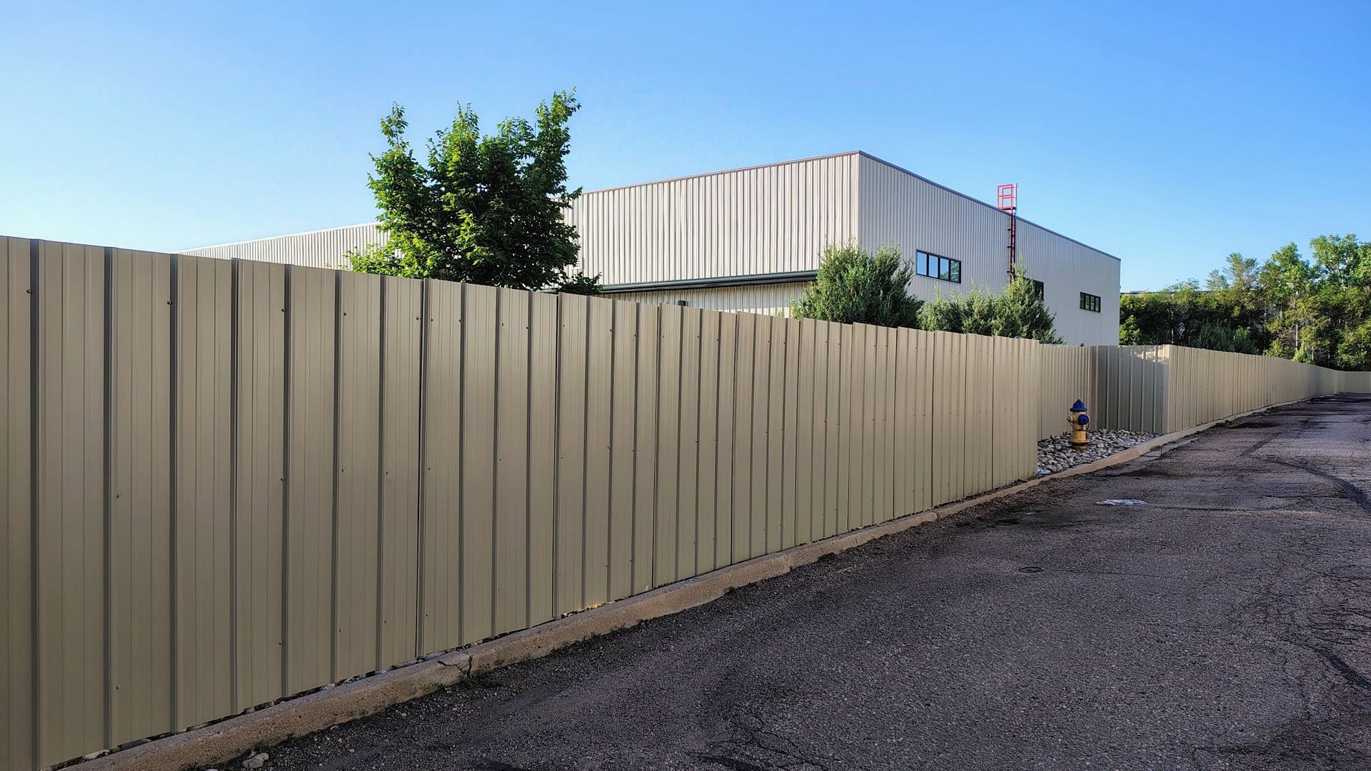 Long beige fence along a gravel road beside a low industrial building under a blue sky