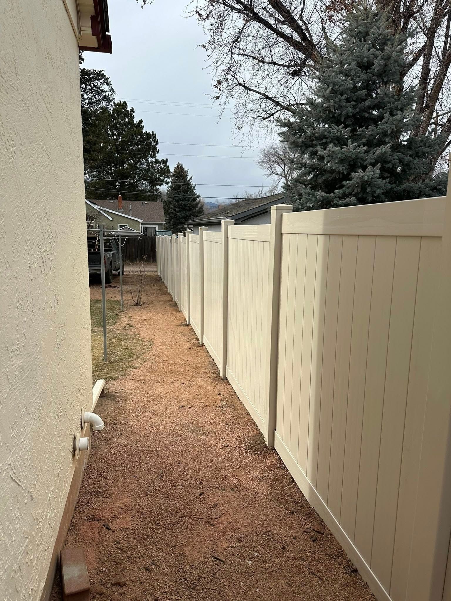 Narrow gravel side yard with a beige privacy fence and tall trees along a house wall