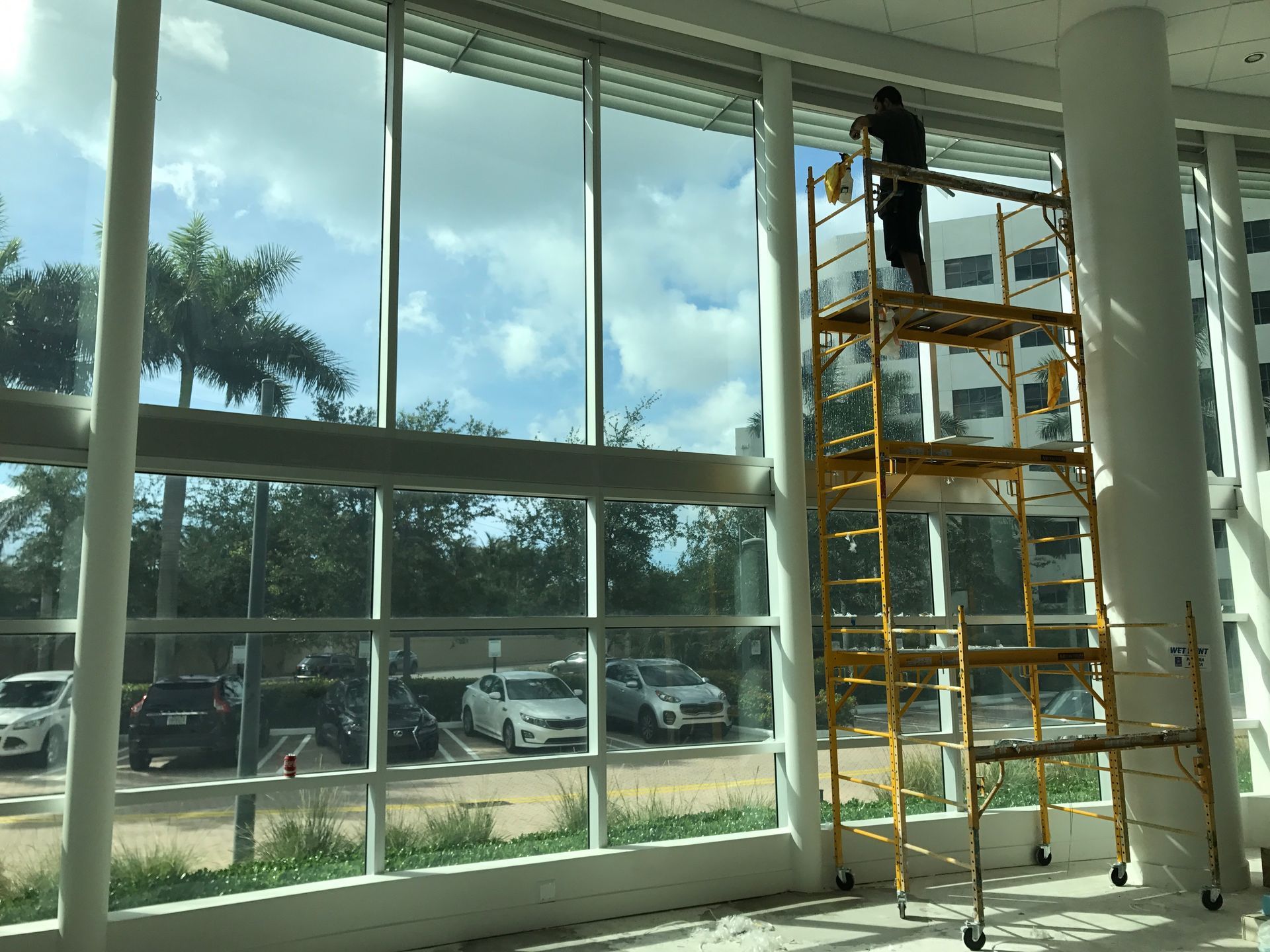Person cleaning windows on scaffolding; building interior with large windows overlooking parking lot.