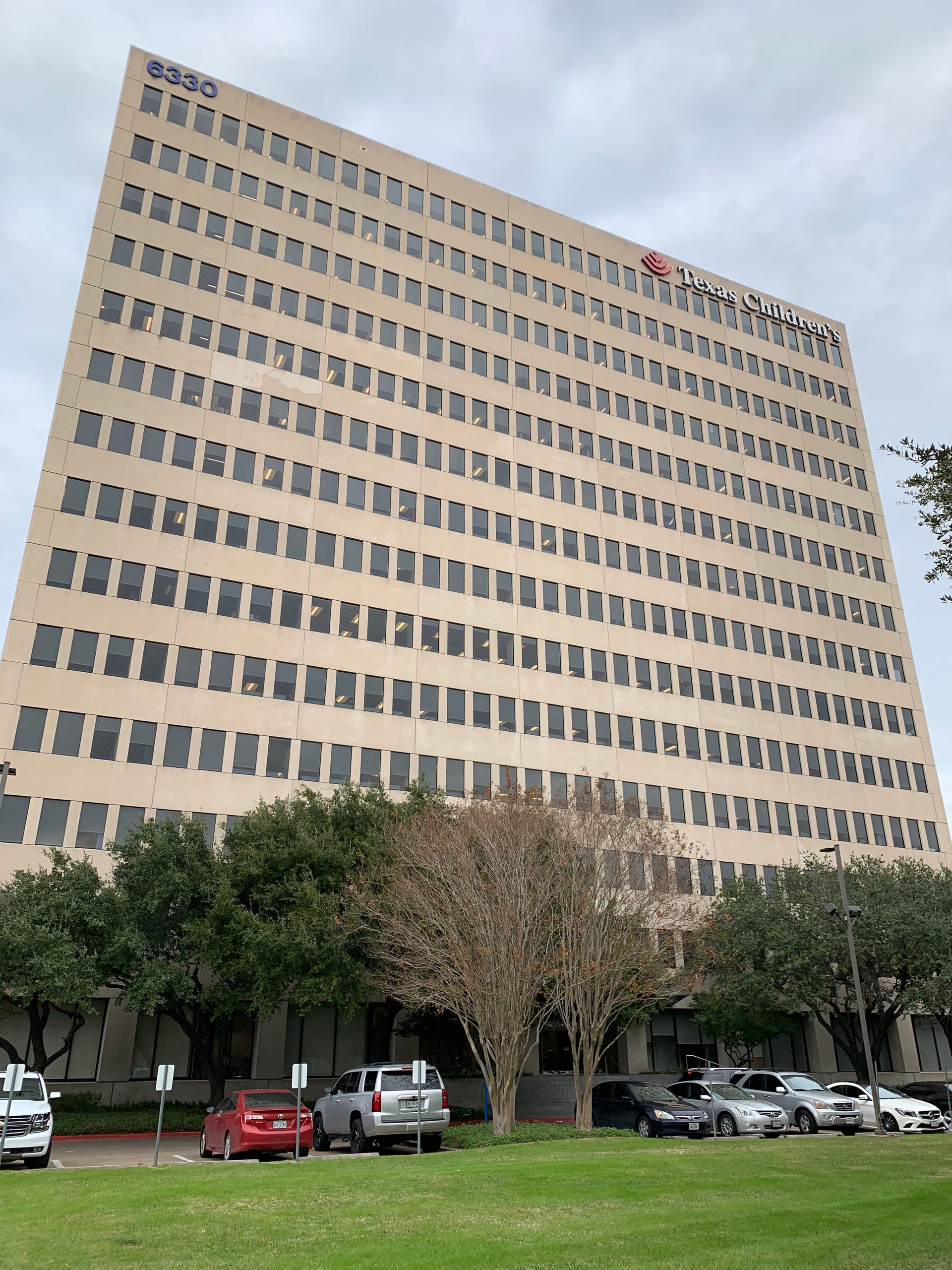 Tall office building with many windows, parked cars in front, and a cloudy sky.