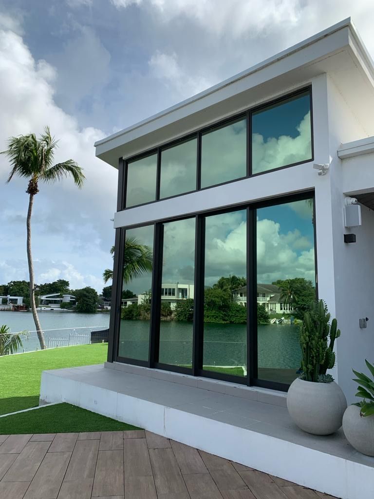 Modern white house with large windows reflecting sky and water; palm tree and lawn in front.