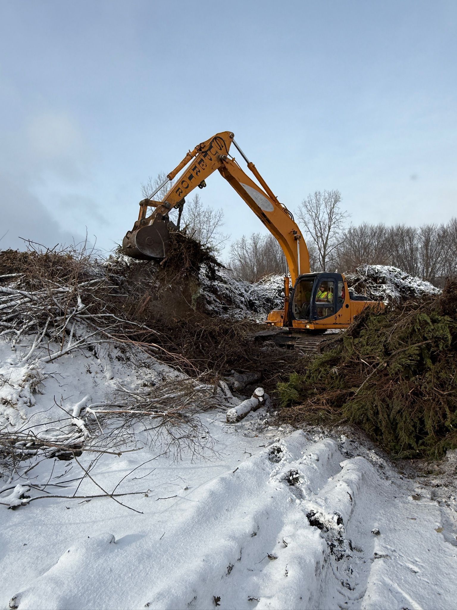 Yellow excavator clearing snow-covered brush on a hillside under a cloudy sky