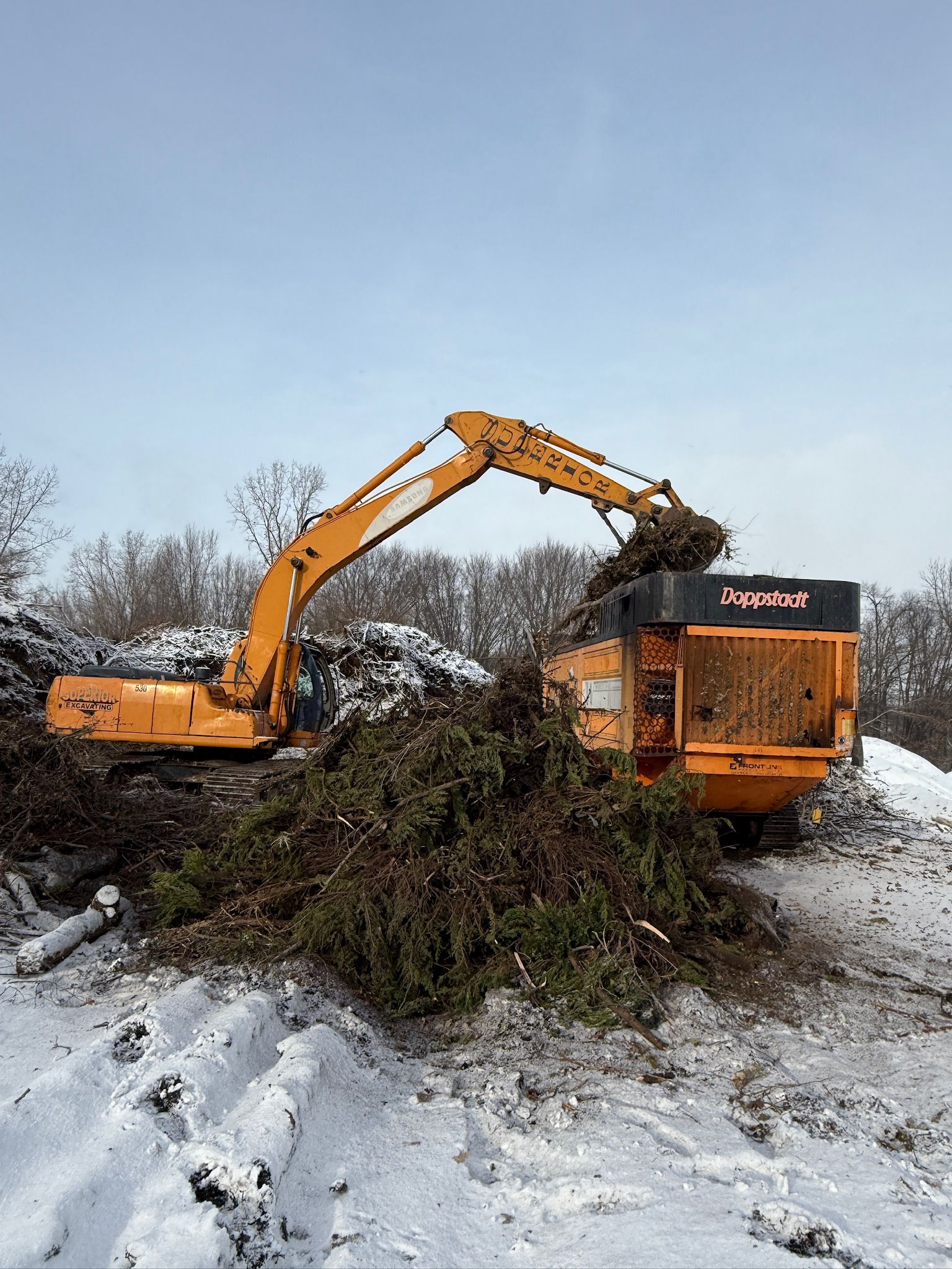 Orange excavator loading brush into a wood chipper in a snowy field
