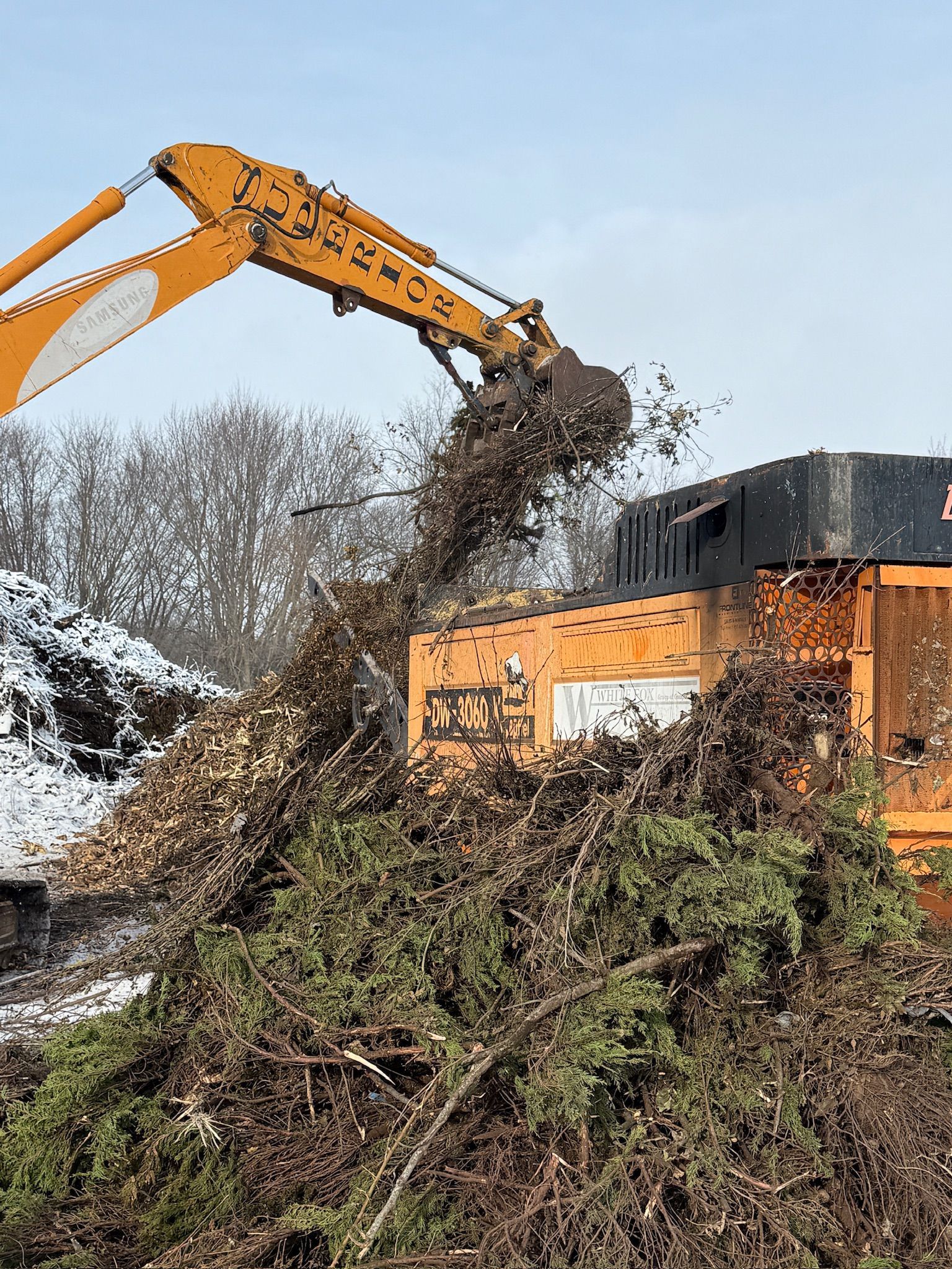 Yellow excavator dumping debris into a brown wood chip pile beside an orange industrial machine outdoors.