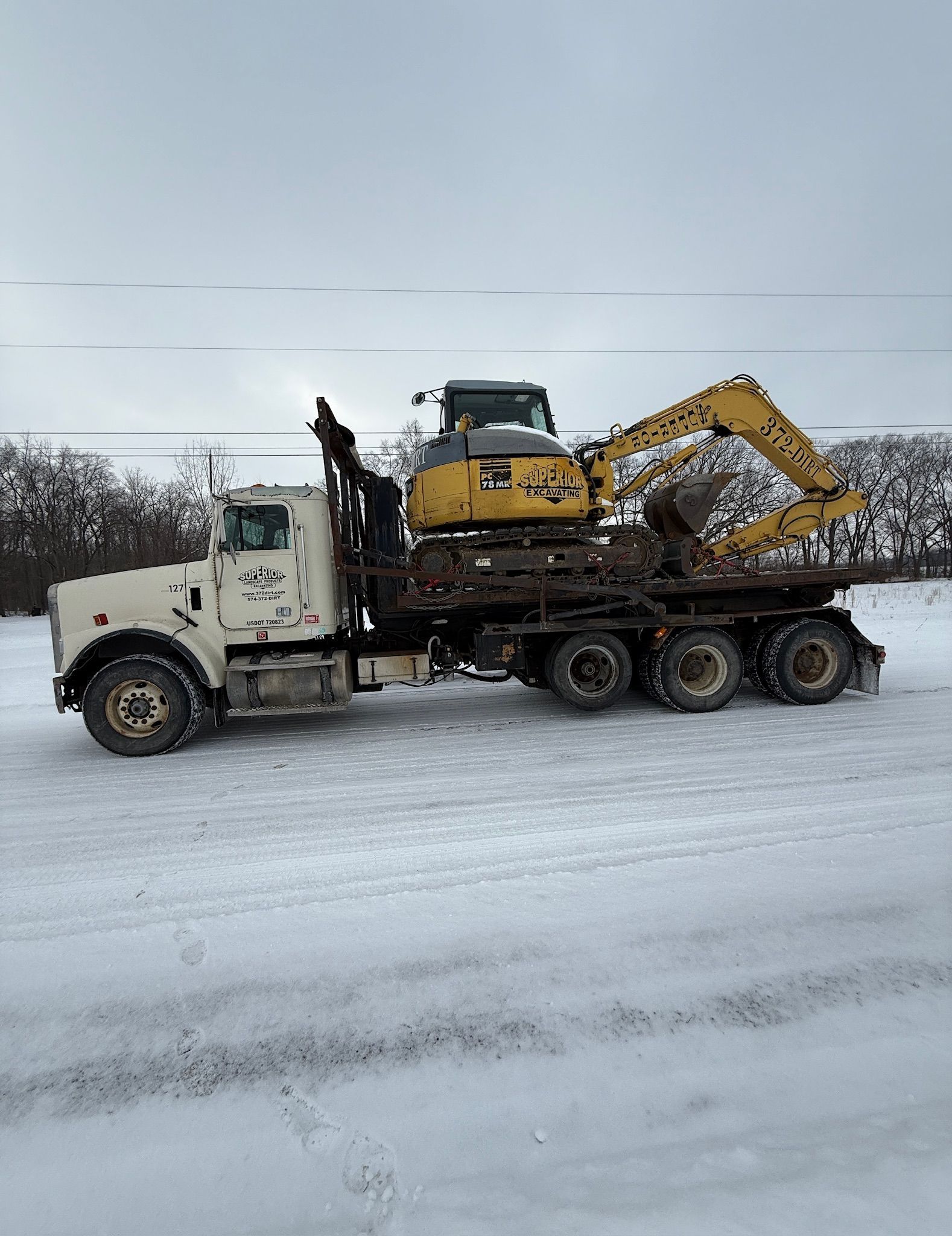 White truck hauling a yellow excavator on a snowy road under an overcast sky