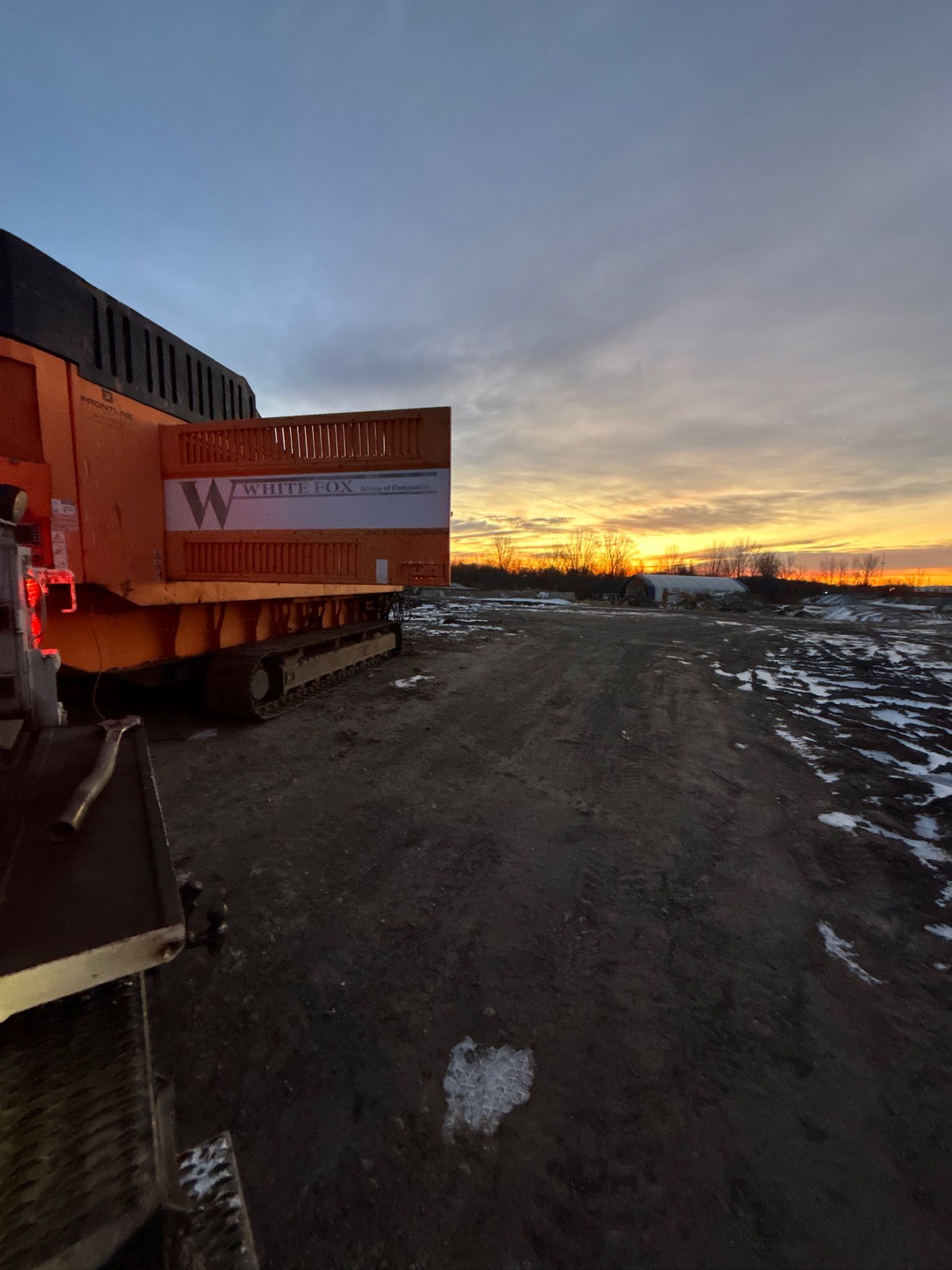 Orange truck parked on a gravel lot at sunset, with snow patches and a glowing horizon.