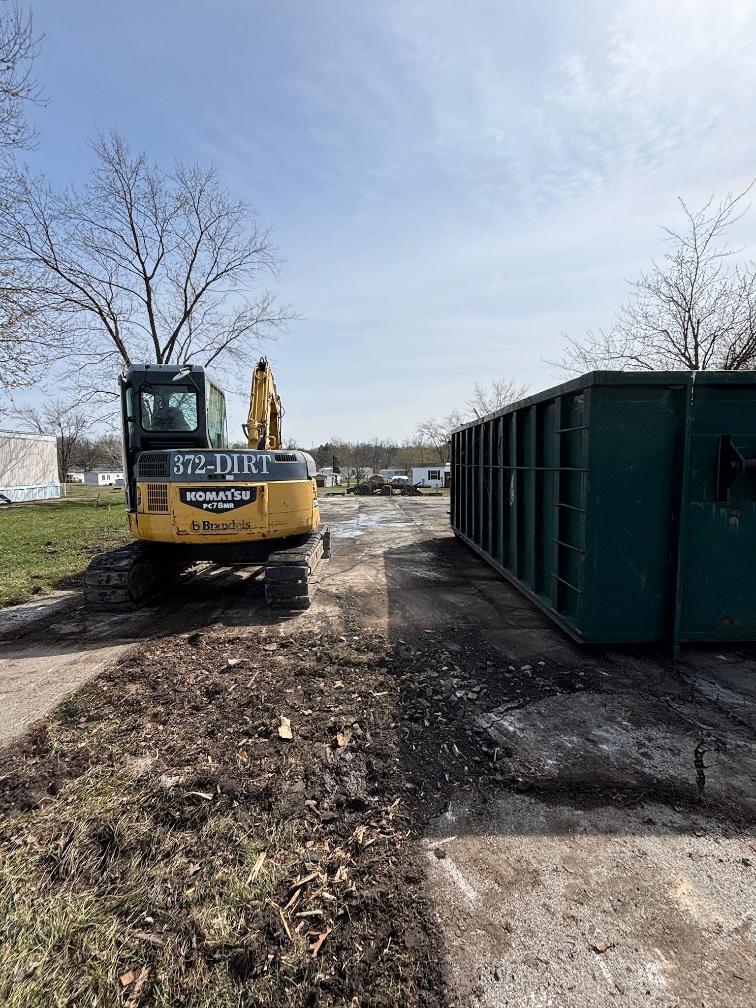 Yellow excavator beside a large green dumpster on a gravel lot under a clear sky