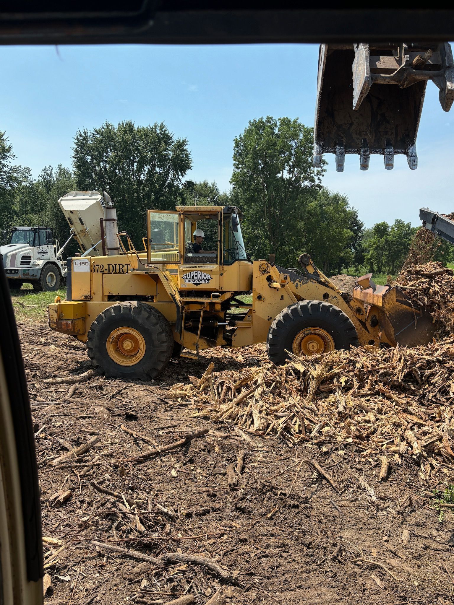 Yellow front loader pushing wood chips at a work site, with trees and a truck in the background