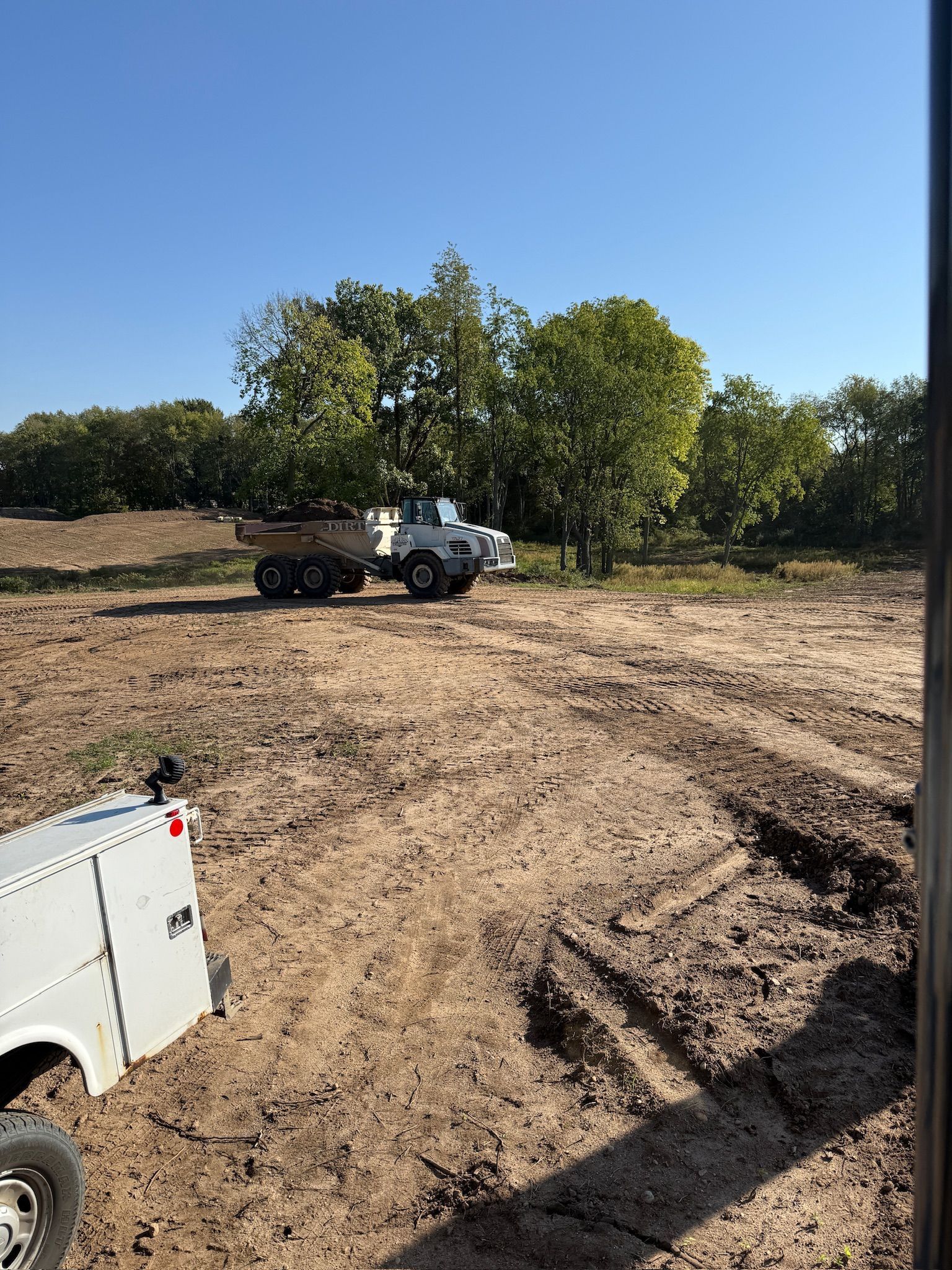 Pickup truck on a dirt field near trees under a clear blue sky, with part of a white truck in the foreground