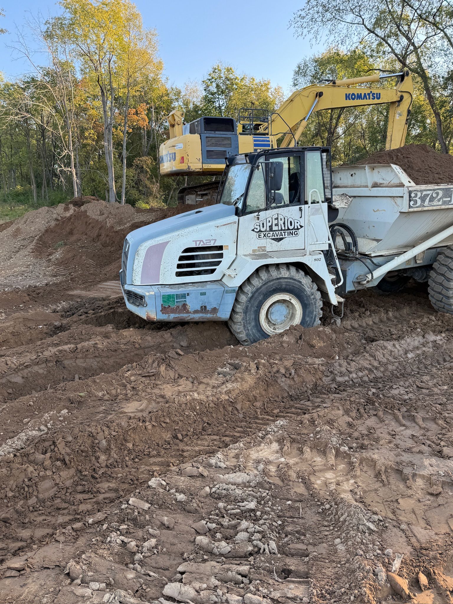 Dump truck and excavator on a muddy construction site with trees in the background