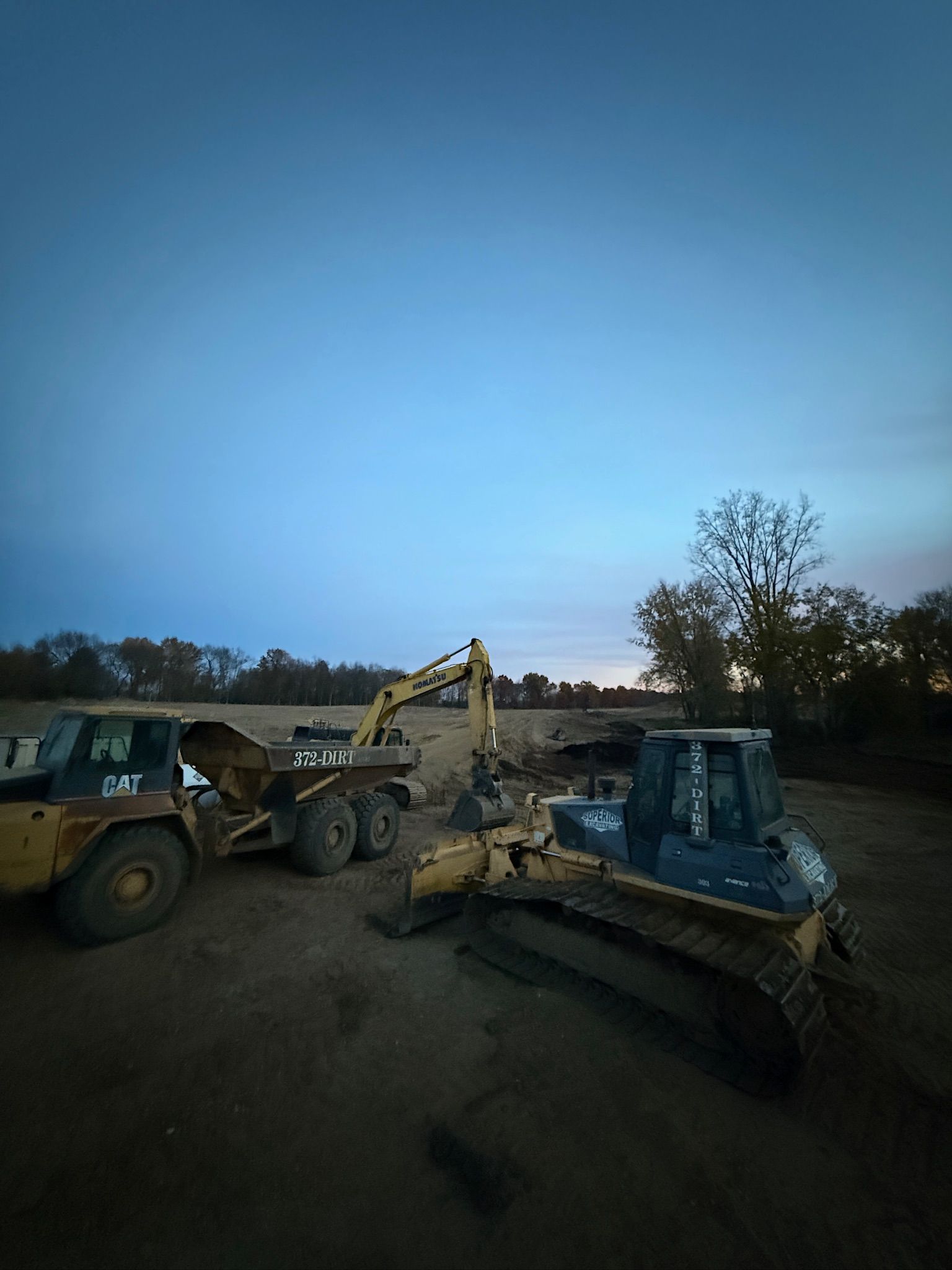 Two construction vehicles parked in a gravel lot at dusk, with a blue sky and trees in the background