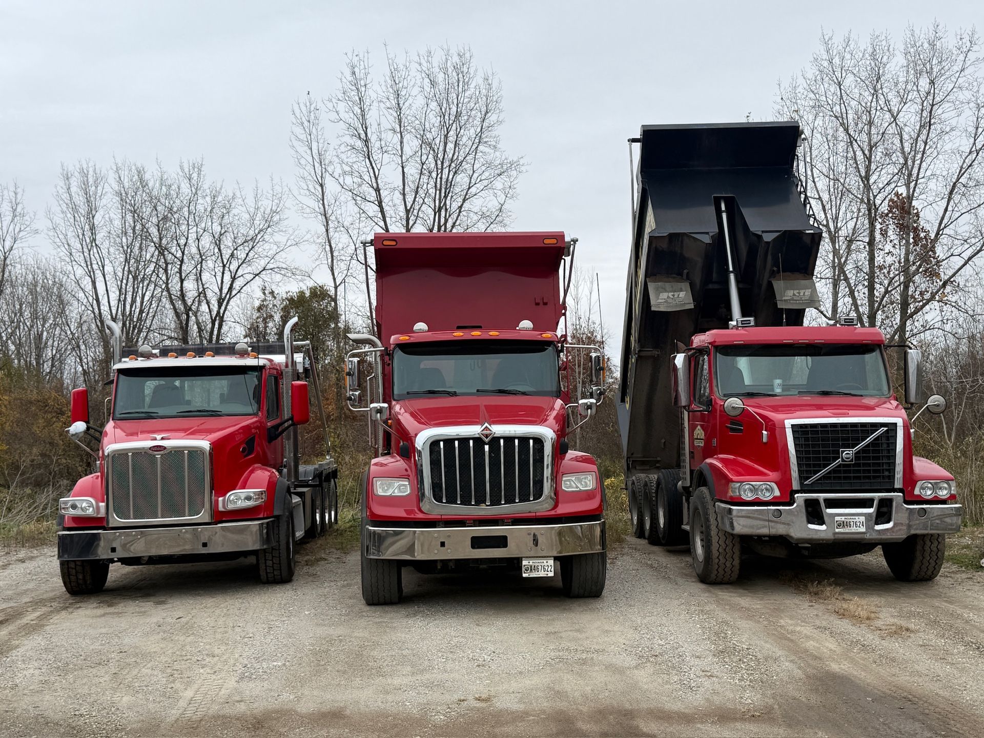 Three red dump trucks parked side by side on a gravel lot with bare trees behind them.