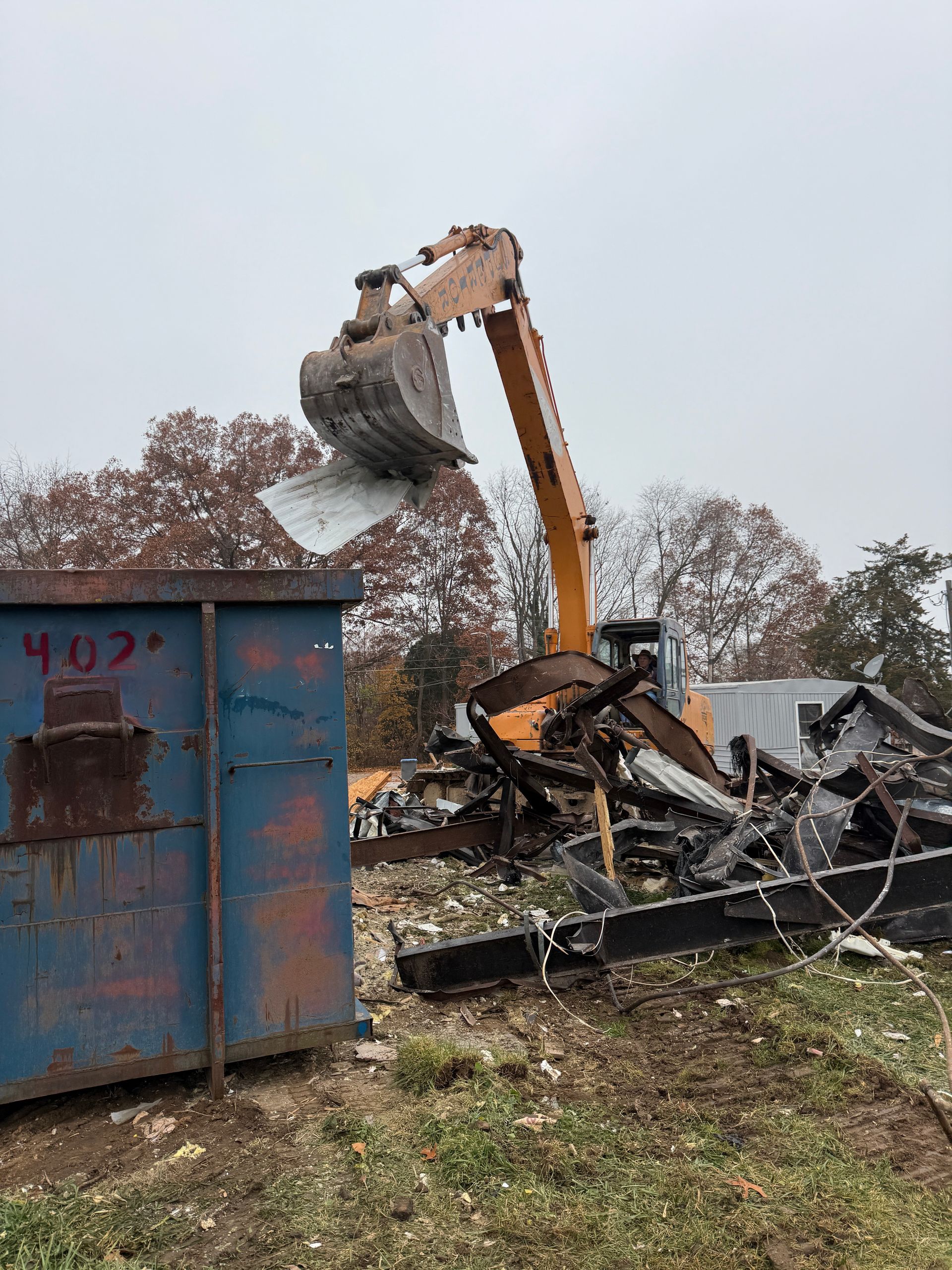 Excavator demolishing debris beside a blue dumpster on a muddy lot under an overcast sky