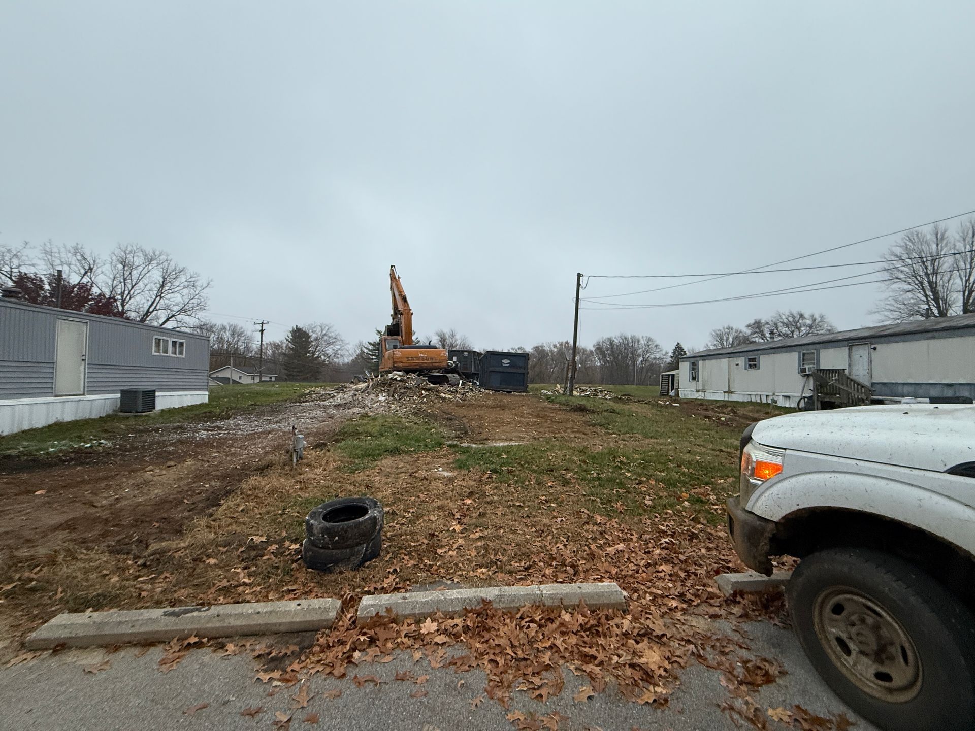 Overcast vacant lot with an excavator, scattered debris, and mobile homes on both sides