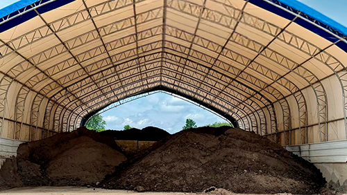 Large piles of dark soil under a beige open-sided agricultural storage tent