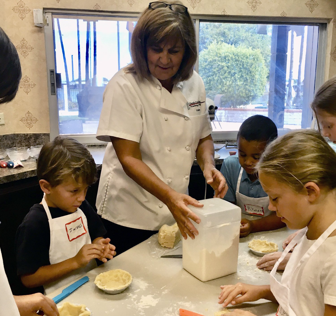 A woman in a chef 's coat is teaching a group of children how to bake