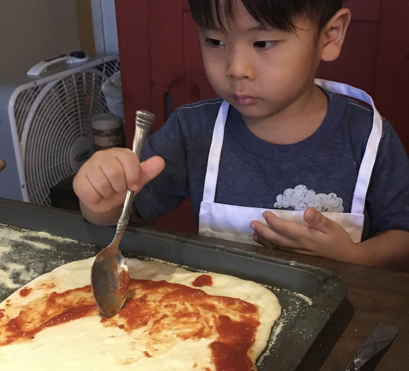A young boy is putting sauce on a pizza with a spoon