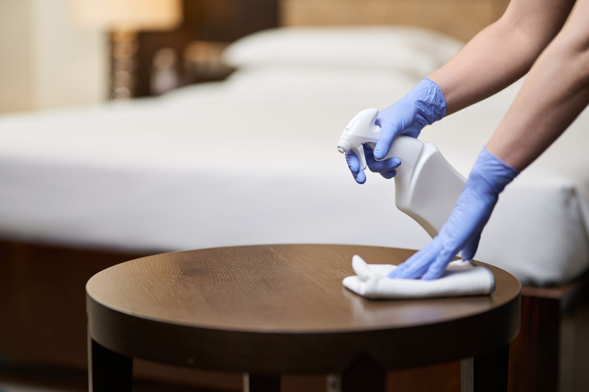 Persona che indossa guanti blu mentre disinfetta un tavolino marrone in una stanza d'albergo.