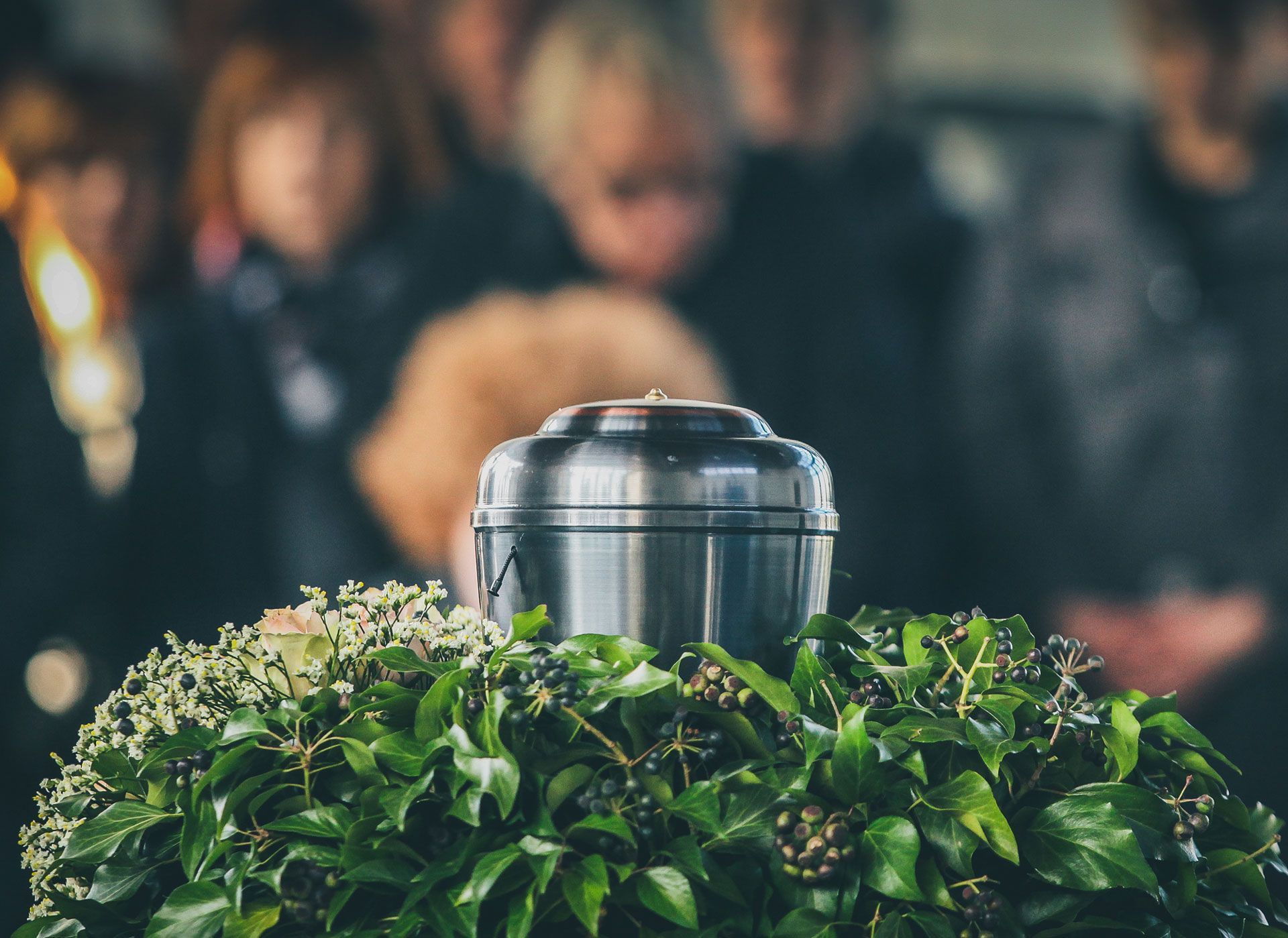 Funeral service with an urn on a floral wreath; mourners in the background, heads bowed in remembrance.
